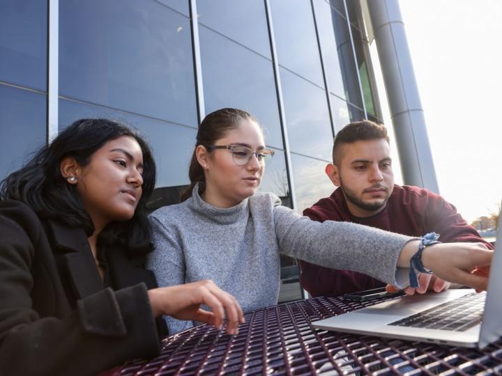 三个学生围坐在户外野餐桌旁的笔记本电脑前讨论课程